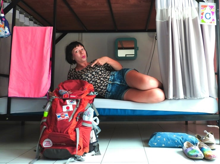 Alice reclining on a hostel bunk beside a red backpack covered in flag stickers, shoes on the floor.