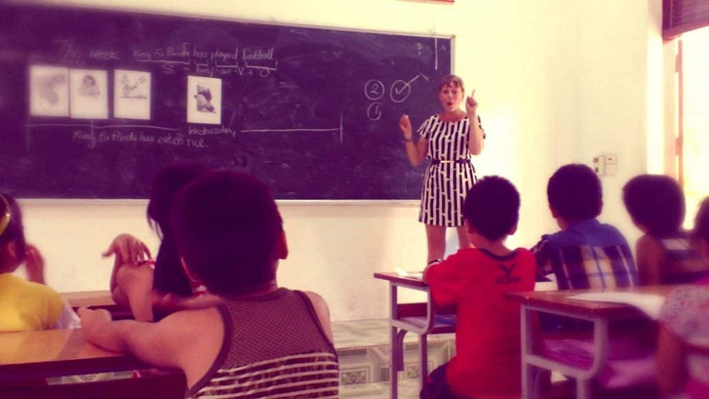 Alice teaching English at a blackboard to a classroom of children seated at desks.