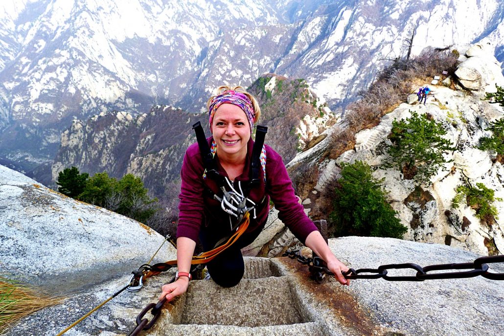 Alice in a purple top smiling at the top of a steep rock stairway with chains on Mount Hua, China.