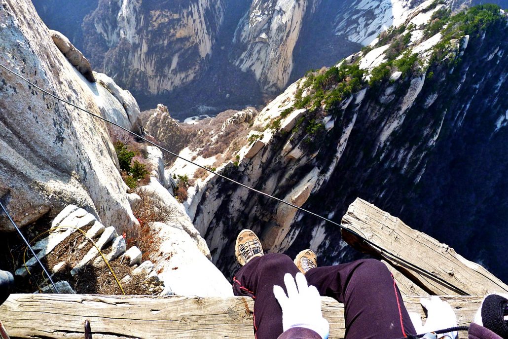 View down past a climber's boots on the wooden plank walk over a sheer drop on Mount Hua.