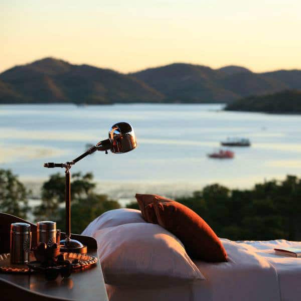 A hotel bed with orange cushions and a reading lamp overlooking Coron bay and hills at sunset.