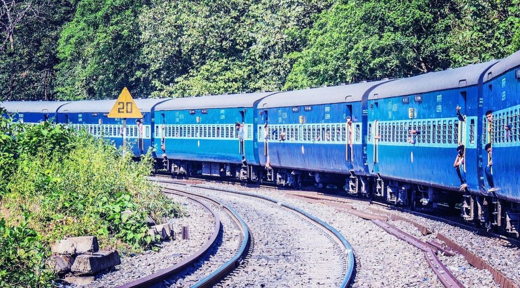 Blue Indian passenger train curving along the tracks past green trees with people at the windows