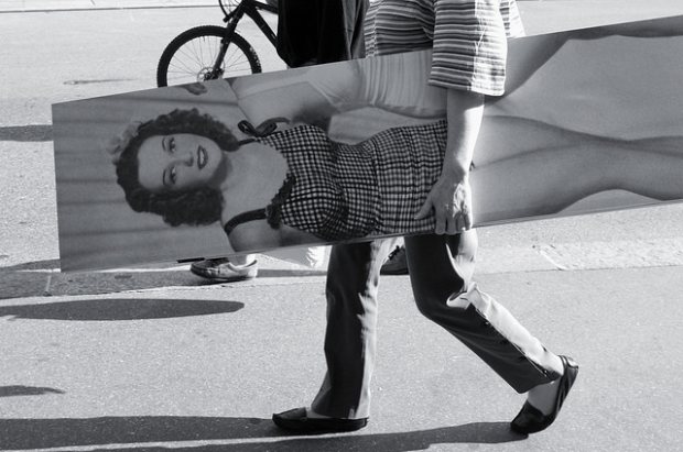 Black and white shot of a person carrying a large printed portrait of a woman across a street