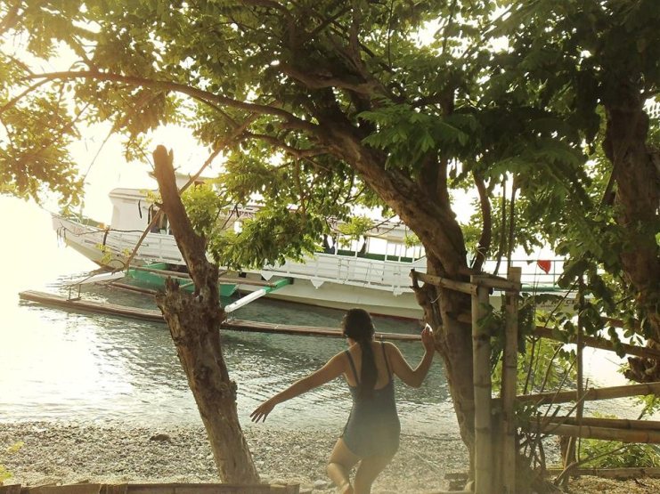 Woman stepping down toward a white outrigger boat moored at the shore under a tree at sunset