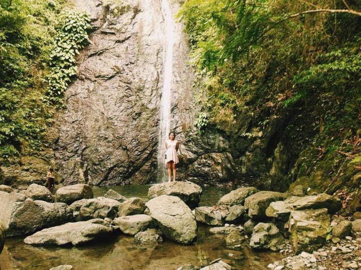 Alice in a white dress standing on a boulder in front of a tall jungle waterfall in the Philippines