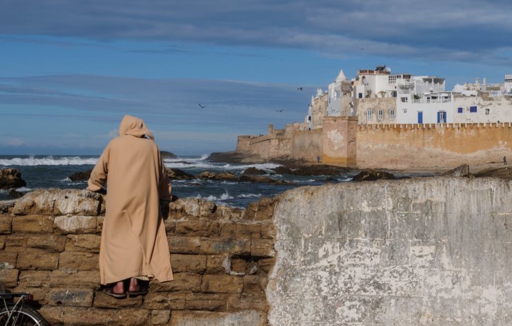 Person in a hooded robe overlooking the sea and the white ramparts of Essaouira, Morocco.