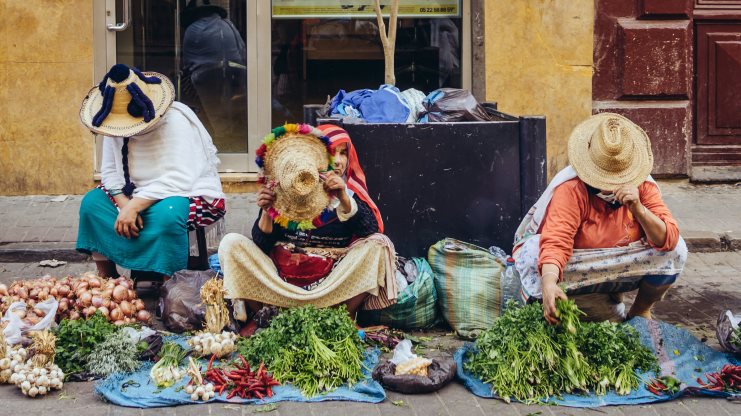Three women in straw hats selling herbs and vegetables on a Moroccan street.