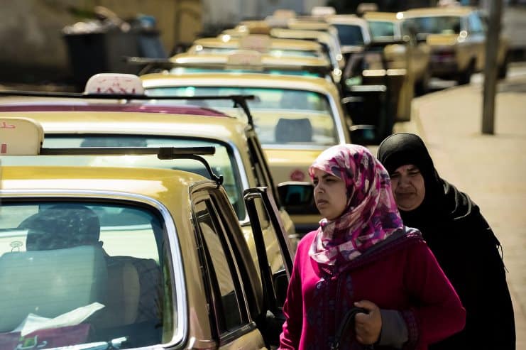 Two women in headscarves walking past a line of parked taxis on a Moroccan street.