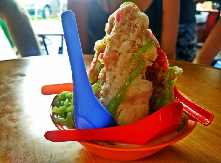 Mound of ais kacang shaved ice with green and red syrup in an orange dish.