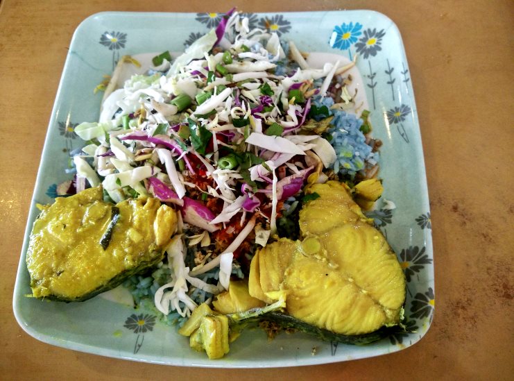 Plate of blue nasi kerabu rice topped with fried fish, herbs and shredded salad.