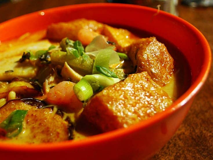 Red bowl of curry laksa with fried tofu puffs, prawns and spring onion.