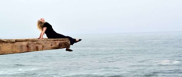 Alice in black reclining on the end of a wooden plank jutting out over the sea.
