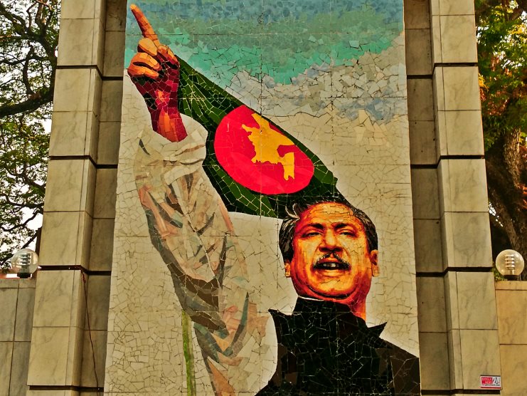 Tiled mosaic monument of a man pointing skyward beside the Bangladesh flag.
