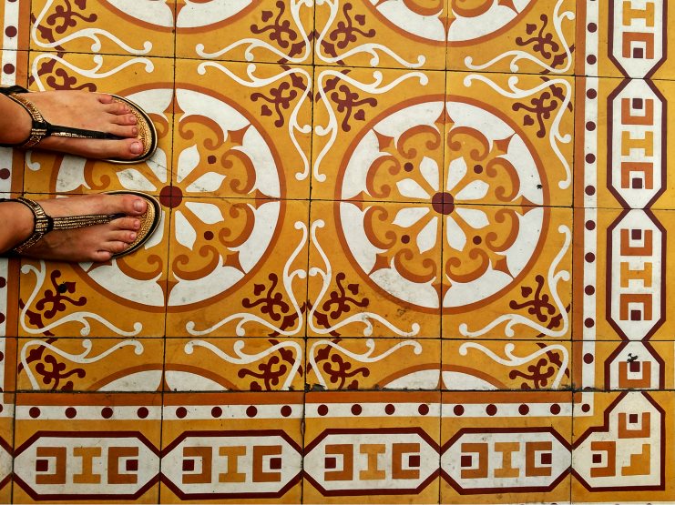 Bare feet in jewelled sandals standing on ornate orange and white patterned floor tiles.