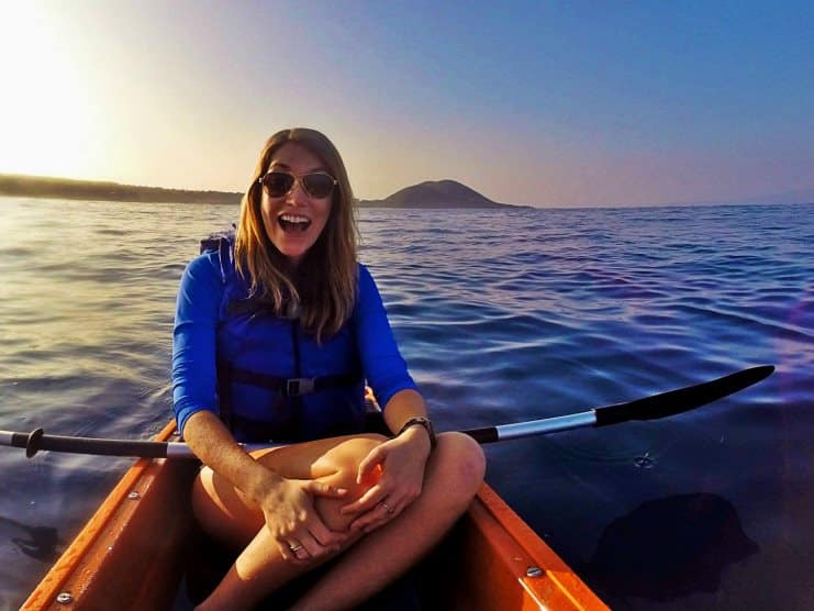 Woman in a blue life jacket laughing while paddling a kayak on calm sea at sunset.