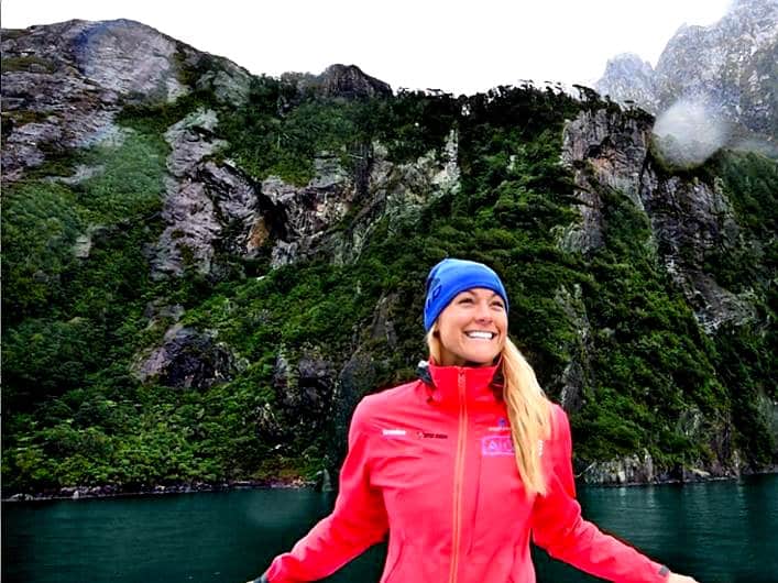 Woman in a red jacket and blue beanie smiling in front of a green fjord cliff.
