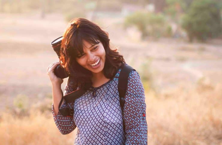 Woman laughing outdoors, holding a camera over her shoulder with a backpack on.