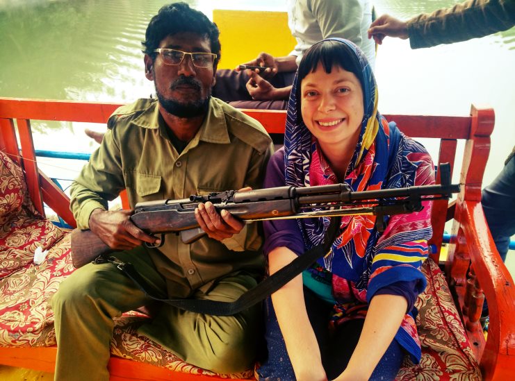 Alice sits beside an armed guard holding a rifle on a boat in the Sundarbans, wearing a printed headscarf.