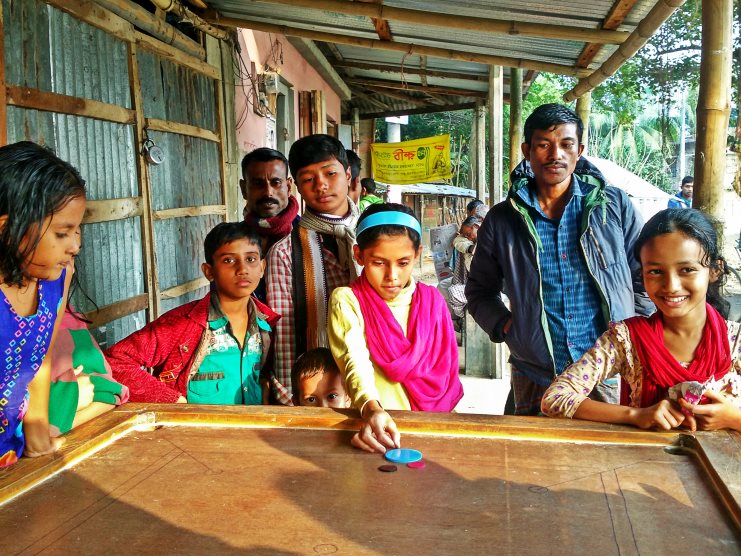 Children and men gather around a carrom board as a girl in yellow flicks a striker.