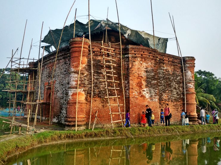 The Chunakhola Mosque under bamboo scaffolding, reflected in a pond, with visitors alongside.