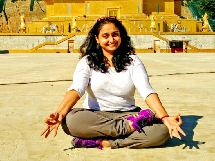 Woman sitting cross-legged and smiling on sand in front of a golden temple complex.