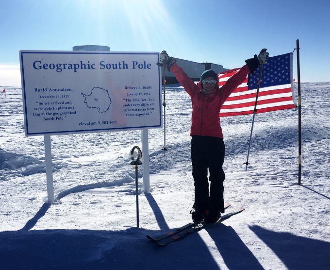 Woman on skis raising ski poles beside the Geographic South Pole sign and a US flag on the ice.
