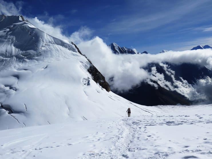 Lone climber with a pack crossing a snowfield towards high Himalayan peaks and clouds.
