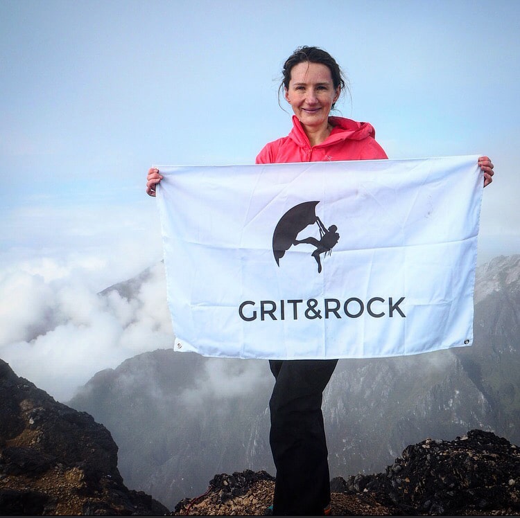 Woman in a red jacket holding a white Grit&Rock flag on a rocky mountain summit above the clouds.