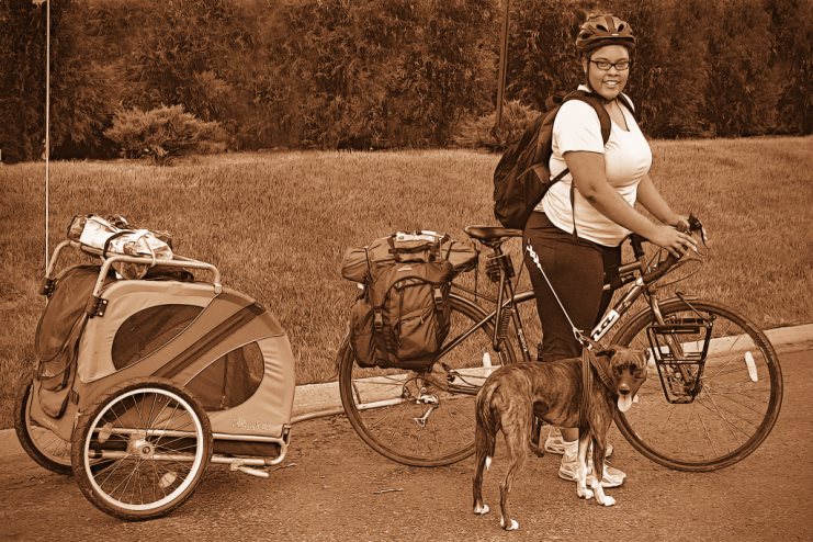 Sepia-toned shot of a woman with a loaded touring bicycle, a trailer of gear and a dog.
