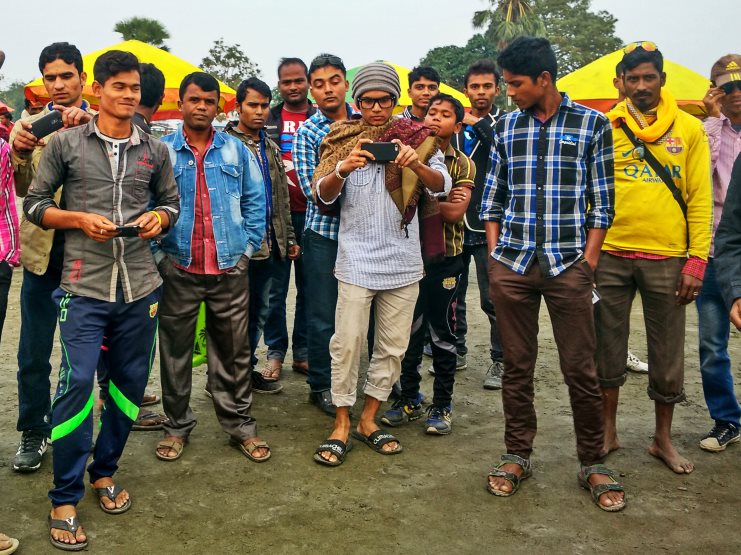 A crowd of young men gather to photograph on their phones in an outdoor Bangladesh setting.