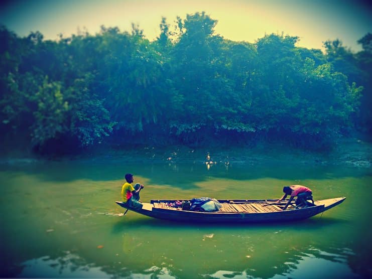 Two men in a wooden boat on a still green river fringed by mangrove forest in the Sundarbans.
