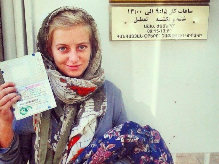 Woman in a headscarf holding up a passport outside an office with Armenian and Farsi signage.