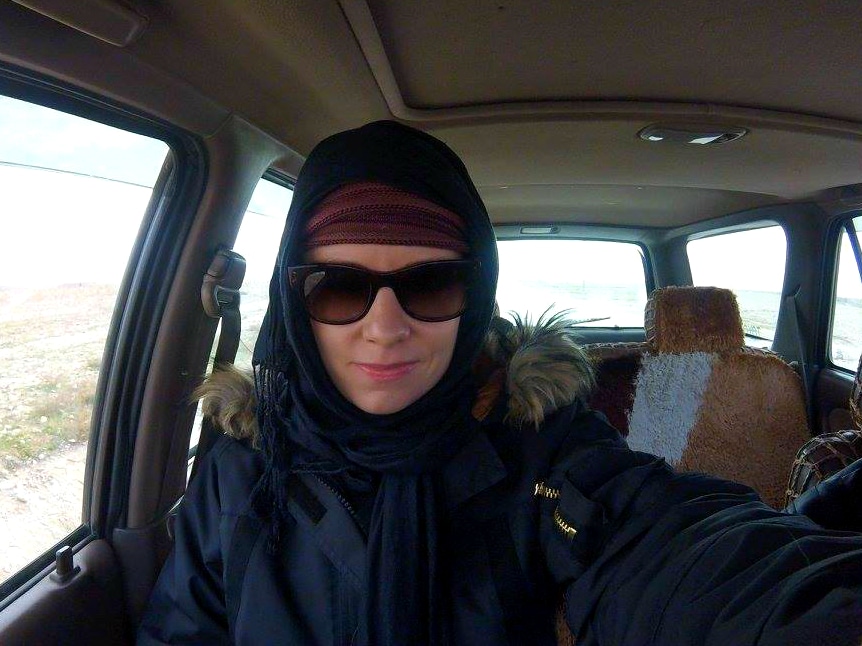 Alice in a hooded coat, headscarf and sunglasses taking a selfie inside a car in a bleak landscape.