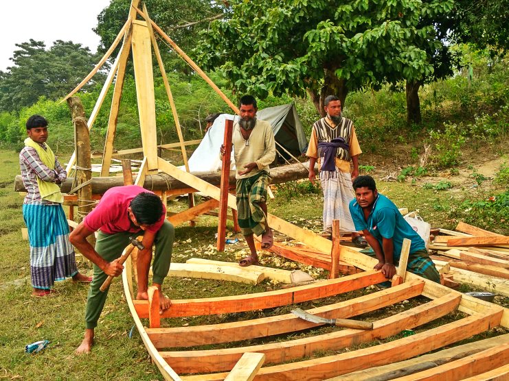 Men build a wooden fishing boat from fresh timber planks on grass near Kuakata.