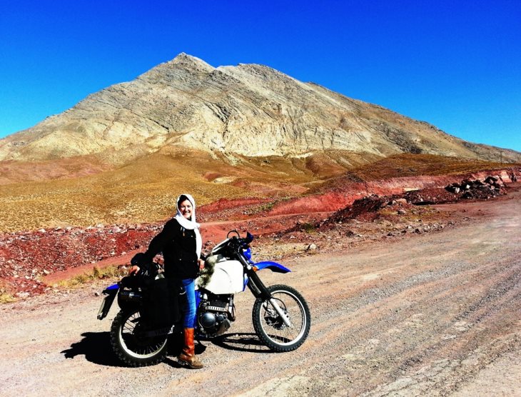 Woman in a headscarf beside a blue motorbike on a desert road towards Yazd, Iran, a bare peak behind.