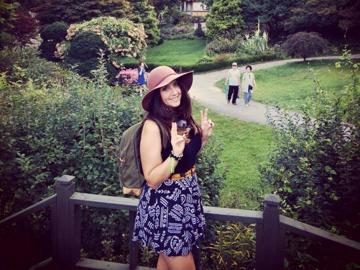 Woman in a wide-brimmed hat and backpack giving a peace sign in a green park.