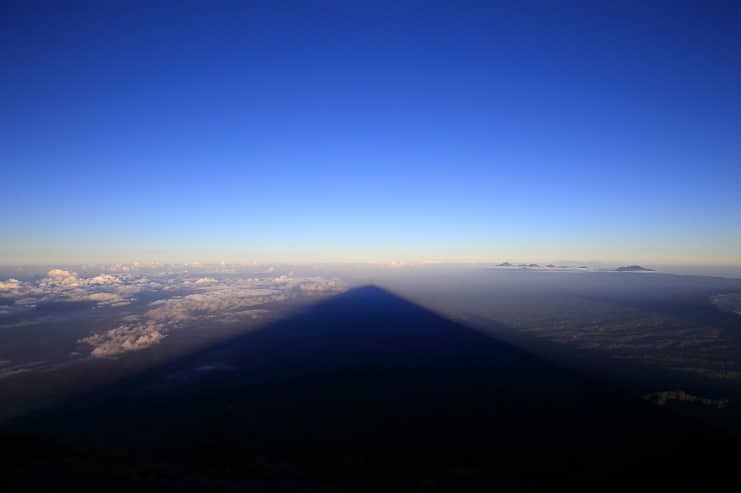 Triangular shadow of a volcano cast across clouds far below, seen from the summit at dawn.