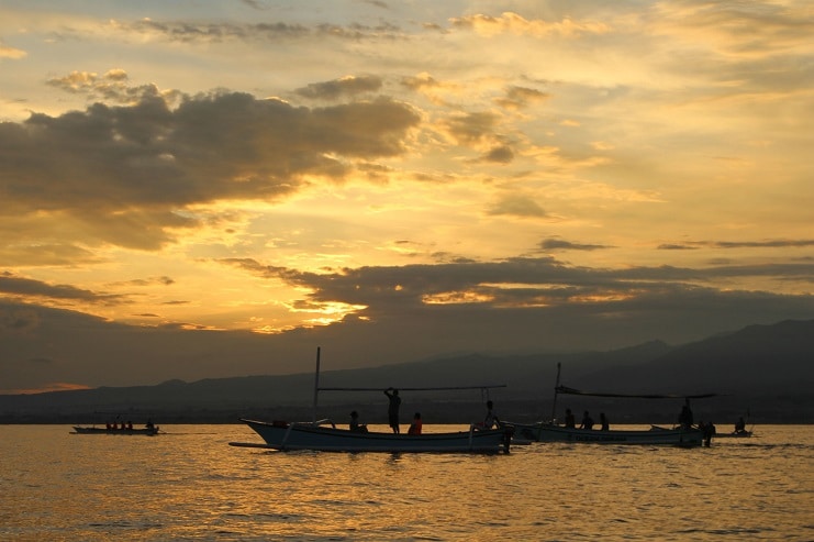 Traditional outrigger fishing boats on calm water at sunset with mountains behind in Bali.