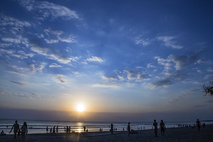 People walking on the beach as the sun sets over the sea under a wide blue sky in Bali.