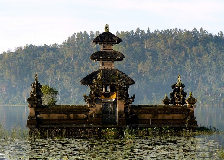 Stone water temple with tiered shrines standing on a still lake, forested hills behind, in Bali.
