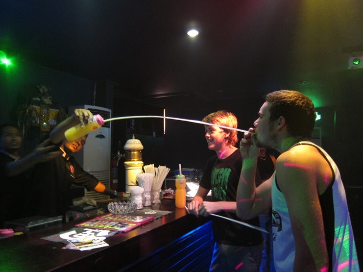Bartender pouring a drink into the mouth of a young man at a neon-lit bar.