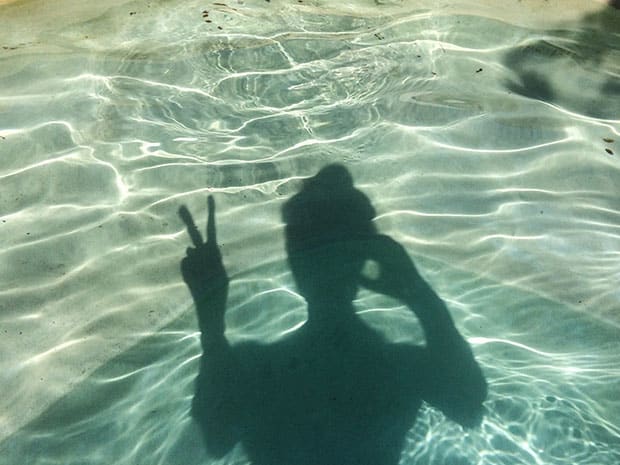 Shadow of a person making a peace sign, cast on the rippling floor of a clear swimming pool.