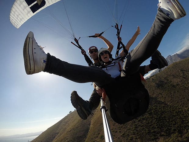 Two people tandem paragliding above green hills, legs kicked out, laughing at a selfie stick camera.
