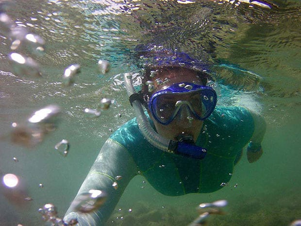 A snorkeler in a green wetsuit and blue mask swims underwater surrounded by bubbles.