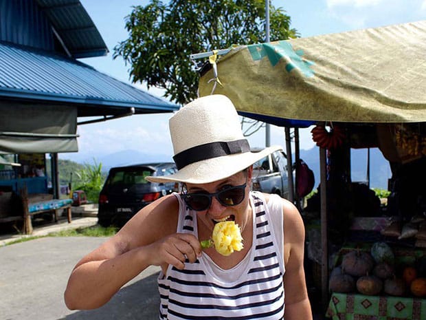 Woman in a straw hat and striped vest eating a slice of fresh pineapple at a roadside fruit stall.