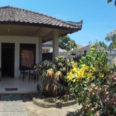 Bali guesthouse with tiled roof and a covered porch beside tropical shrubs and yellow flowers.