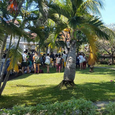 Travellers gathered with backpacks on a lawn among coconut palms at Bayu Mantra Hotel in Bali.