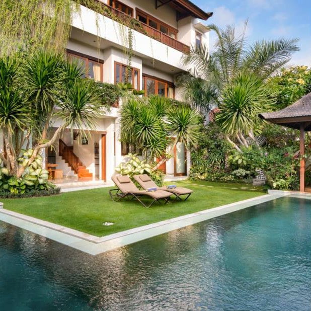 Bali villa with two sun loungers on a lawn beside an L-shaped pool and palm trees.