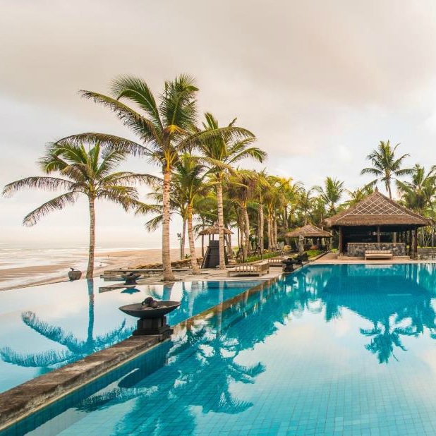 Beachfront infinity pool lined with palm trees and a thatched pavilion at Legian Beach, Bali.