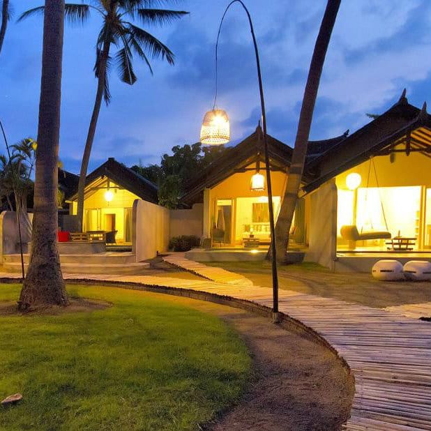Thatched-roof villas lit at dusk along a bamboo walkway at Lillin Lovina Beach Hotel, Bali.